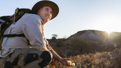 Ecologist Graeme Finlayson crouches down at Evelyn Downs, reaching out to cup a small plant's fluffy seed heads in his hand while looking off into the distance.