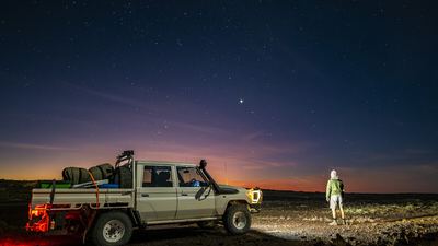 Under a spectacular, star-dusted evening sky, a person stands next to a work ute loaded up with gear, their back to the camera, looking towards a horizon streaked with sunset colours.