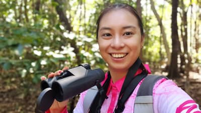 Bush Heritage volunteer Eva You, a young Asian-Australian woman, stands outdoors holding binoculars and smiling.