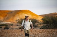 Ecologist Graeme Finlayson strides towards the camera against a backdrop of Evelyn Downs's low, golden-yellow hills.