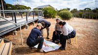 A small group of four people gather in a circle, looking at a map on the ground out the front of the Red Moort Field Station.