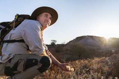 Ecologist Graeme Finlayson crouches down at Evelyn Downs, reaching out to cup a small plant's fluffy seed heads in his hand while looking off into the distance.