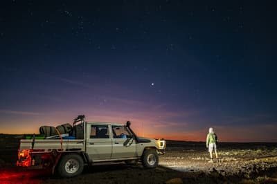 Under a spectacular, star-dusted evening sky, a person stands next to a work ute loaded up with gear, their back to the camera, looking towards a horizon streaked with sunset colours.