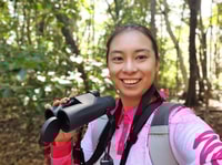 Bush Heritage volunteer Eva You, a young Asian-Australian woman, stands outdoors holding binoculars and smiling.