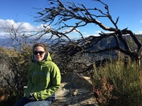 Bush Heritage volunteer Georgia McManus, a young woman in a green parka, sits on a rock at the top of a mountain.